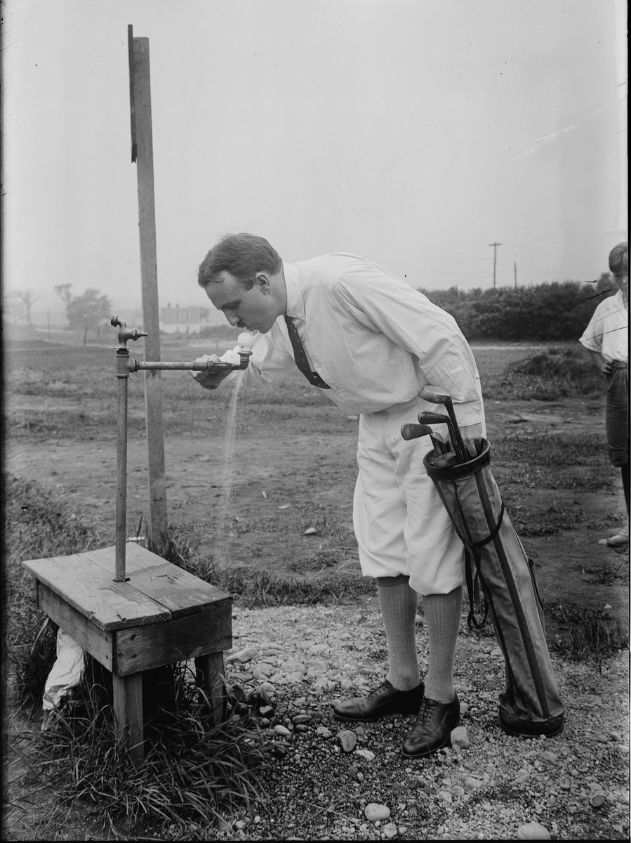 Photographie ancienne d'un golfeur qui boit à une fontaine sur le parcours, son sac à côté